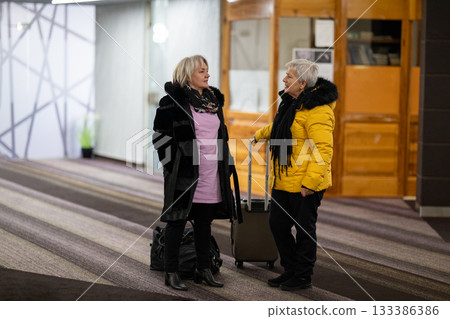 Two older women dressed in winter clothing standing in a hotel lobby and having a friendly conversation next to their luggage. Concept of travel, friendship, and leisure lifestyle for seniors. 133386386
