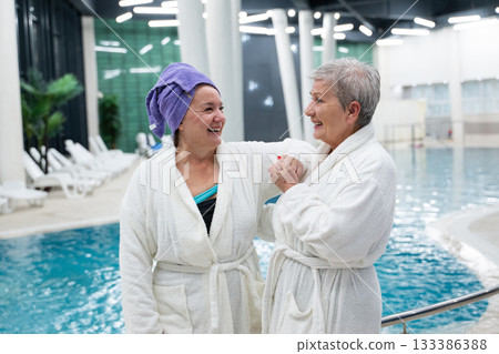 Two cheerful senior women in white bathrobes laughing and relaxing together at an indoor swimming pool. Concept of friendship, wellness, spa lifestyle, and healthy aging. 133386388