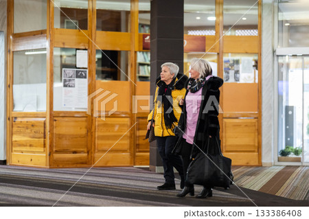 Two older women dressed in winter clothing standing in a hotel lobby and having a friendly conversation next to their luggage. Concept of travel, friendship, and leisure lifestyle for seniors. Two older women dressed in winter clothing standing in a hotel lobby and having a friendly conversation next to their luggage. Concept of travel, friendship, and leisure lifestyle for seniors. 133386408