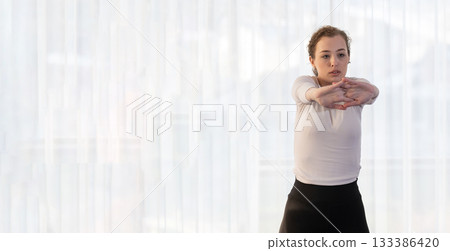 Focused young woman in a white shirt performing a stretching exercise indoors. Concept of wellness, health, flexibility, mindfulness, and active lifestyle. 133386420