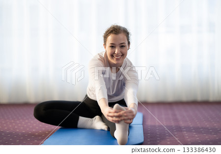 Focused woman stretching her leg while sitting on a yoga mat in a bright indoor space. Concept of wellness, flexibility, mindfulness, and healthy lifestyle through exercise and relaxation. Focused woman stretching her leg while sitting on a yoga mat in a bright indoor space. Concept of wellness, flexibility, mindfulness, and healthy lifestyle through exercise and relaxation. 133386430