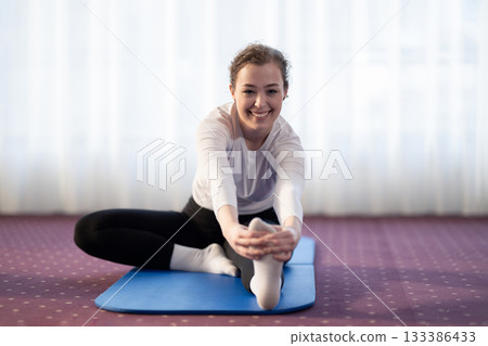 Focused woman stretching her leg while sitting on a yoga mat in a bright indoor space. Concept of wellness, flexibility, mindfulness, and healthy lifestyle through exercise and relaxation. 133386433