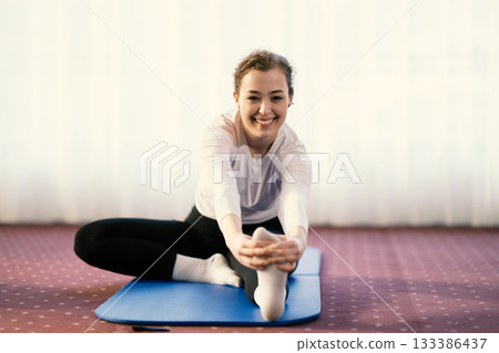 Focused woman stretching her leg while sitting on a yoga mat in a bright indoor space. Concept of wellness, flexibility, mindfulness, and healthy lifestyle through exercise and relaxation. Focused woman stretching her leg while sitting on a yoga mat in a bright indoor space. Concept of wellness, flexibility, mindfulness, and healthy lifestyle through exercise and relaxation. 133386437