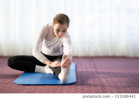 Focused woman stretching her leg while sitting on a yoga mat in a bright indoor space. Concept of wellness, flexibility, mindfulness, and healthy lifestyle through exercise and relaxation. Focused woman stretching her leg while sitting on a yoga mat in a bright indoor space. Concept of wellness, flexibility, mindfulness, and healthy lifestyle through exercise and relaxation. 133386441
