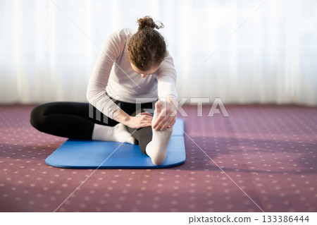 Focused woman stretching her leg while sitting on a yoga mat in a bright indoor space. Concept of wellness, flexibility, mindfulness, and healthy lifestyle through exercise and relaxation. 133386444