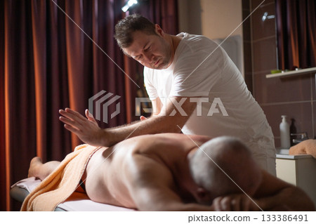 Professional therapist applying deep tissue massage techniques on a male client lying on massage table with towel, therapeutic bodywork for muscle pain relief and relaxation. Professional therapist applying deep tissue massage techniques on a male client lying on massage table with towel, therapeutic bodywork for muscle pain relief and relaxation. 133386491