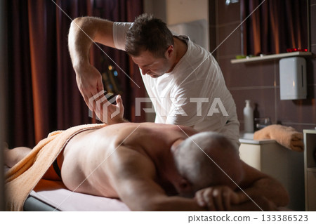 Professional therapist applying deep tissue massage techniques on a male client lying on massage table with towel, therapeutic bodywork for muscle pain relief and relaxation. 133386523