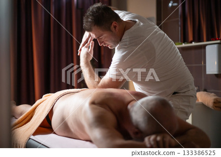 Professional therapist applying deep tissue massage techniques on a male client lying on massage table with towel, therapeutic bodywork for muscle pain relief and relaxation. Professional therapist applying deep tissue massage techniques on a male client lying on massage table with towel, therapeutic bodywork for muscle pain relief and relaxation. 133386525