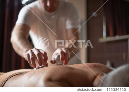 Close up of therapist applying cupping therapy on male clients back during wellness and rehabilitation treatment, traditional alternative medicine for relaxation and pain relief. 133386530
