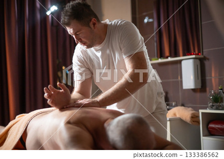 Professional therapist applying deep tissue massage techniques on a male client lying on massage table with towel, therapeutic bodywork for muscle pain relief and relaxation. 133386562