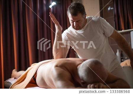 Professional therapist applying deep tissue massage techniques on a male client lying on massage table with towel, therapeutic bodywork for muscle pain relief and relaxation. 133386568