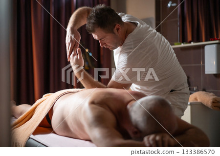 Professional therapist applying deep tissue massage techniques on a male client lying on massage table with towel, therapeutic bodywork for muscle pain relief and relaxation. Professional therapist applying deep tissue massage techniques on a male client lying on massage table with towel, therapeutic bodywork for muscle pain relief and relaxation. 133386570