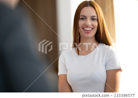 Cheerful young woman smiling and looking at the camera while standing indoors in casual white clothing. Concept of positivity, confidence, happiness, and natural beauty. Cheerful young woman smiling and looking at the camera while standing indoors in casual white clothing. Concept of positivity, confidence, happiness, and natural beauty. 133386577