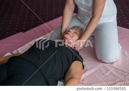 Close up of a physiotherapist holding the head and neck of an older woman lying on a mat during a rehabilitation session. Physiotherapy treatment concept focusing on pain relief, mobility improvement Close up of a physiotherapist holding the head and neck of an older woman lying on a mat during a rehabilitation session. Physiotherapy treatment concept focusing on pain relief, mobility improvement 133386585