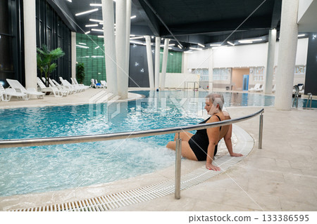 Elderly woman sitting by the edge of a modern indoor spa pool, enjoying the relaxing water jets. Wellness and relaxation concept in a luxurious spa or wellness center. 133386595