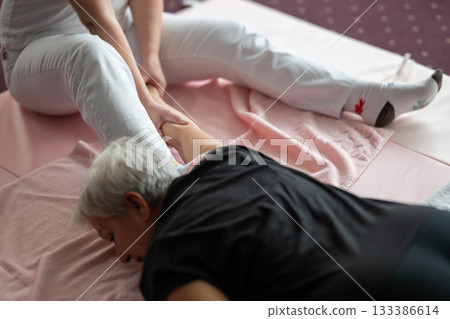 A physiotherapist assisting an older woman with a shoulder and arm stretch while lying on a mat. Rehabilitation and physiotherapy session concept focusing on mobility, flexibility, pain relief, and 133386614
