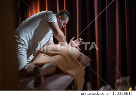 Professional male massage therapist performing deep tissue back massage on an older man lying on a massage table covered with a towel. Relaxing wellness treatment in a modern spa room with warm light Professional male massage therapist performing deep tissue back massage on an older man lying on a massage table covered with a towel. Relaxing wellness treatment in a modern spa room with warm light 133386615