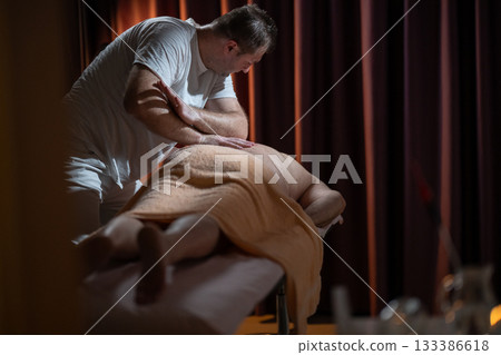 Professional male massage therapist performing deep tissue back massage on an older man lying on a massage table covered with a towel. Relaxing wellness treatment in a modern spa room with warm light Professional male massage therapist performing deep tissue back massage on an older man lying on a massage table covered with a towel. Relaxing wellness treatment in a modern spa room with warm light 133386618