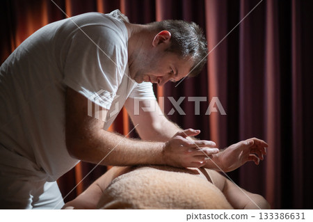 Professional male massage therapist performing deep tissue back massage on an older man lying on a massage table covered with a towel. Relaxing wellness treatment in a modern spa room with warm light Professional male massage therapist performing deep tissue back massage on an older man lying on a massage table covered with a towel. Relaxing wellness treatment in a modern spa room with warm light 133386631
