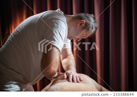 Professional male massage therapist performing deep tissue back massage on an older man lying on a massage table covered with a towel. Relaxing wellness treatment in a modern spa room with warm light 133386644