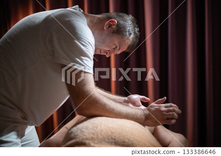 Professional male massage therapist performing deep tissue back massage on an older man lying on a massage table covered with a towel. Relaxing wellness treatment in a modern spa room with warm light Professional male massage therapist performing deep tissue back massage on an older man lying on a massage table covered with a towel. Relaxing wellness treatment in a modern spa room with warm light 133386646