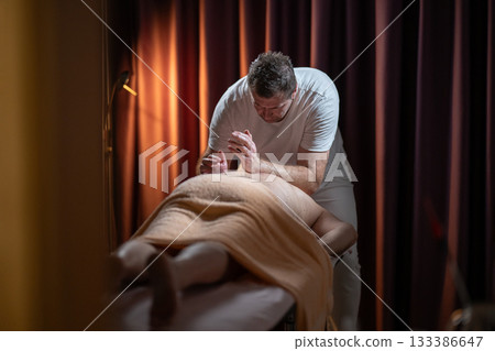Professional male massage therapist performing deep tissue back massage on an older man lying on a massage table covered with a towel. Relaxing wellness treatment in a modern spa room with warm light Professional male massage therapist performing deep tissue back massage on an older man lying on a massage table covered with a towel. Relaxing wellness treatment in a modern spa room with warm light 133386647