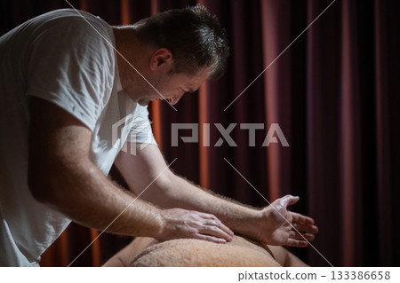 Professional male massage therapist performing deep tissue back massage on an older man lying on a massage table covered with a towel. Relaxing wellness treatment in a modern spa room with warm light Professional male massage therapist performing deep tissue back massage on an older man lying on a massage table covered with a towel. Relaxing wellness treatment in a modern spa room with warm light 133386658