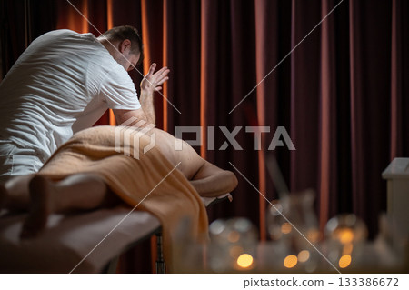 Professional male massage therapist performing deep tissue back massage on an older man lying on a massage table covered with a towel. Relaxing wellness treatment in a modern spa room with warm light Professional male massage therapist performing deep tissue back massage on an older man lying on a massage table covered with a towel. Relaxing wellness treatment in a modern spa room with warm light 133386672
