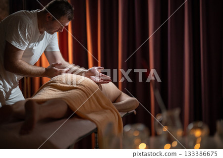 Professional male massage therapist performing deep tissue back massage on an older man lying on a massage table covered with a towel. Relaxing wellness treatment in a modern spa room with warm light Professional male massage therapist performing deep tissue back massage on an older man lying on a massage table covered with a towel. Relaxing wellness treatment in a modern spa room with warm light 133386675