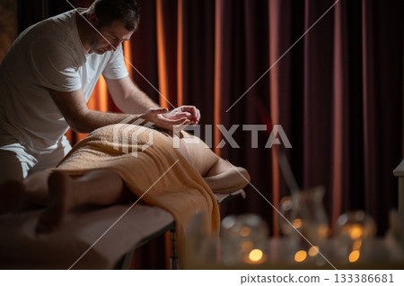 Professional male massage therapist performing deep tissue back massage on an older man lying on a massage table covered with a towel. Relaxing wellness treatment in a modern spa room with warm light 133386681