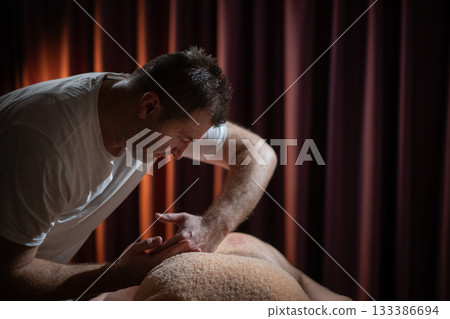Professional male massage therapist performing deep tissue back massage on an older man lying on a massage table covered with a towel. Relaxing wellness treatment in a modern spa room with warm light Professional male massage therapist performing deep tissue back massage on an older man lying on a massage table covered with a towel. Relaxing wellness treatment in a modern spa room with warm light 133386694