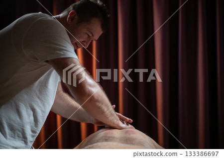 Professional male massage therapist performing deep tissue back massage on an older man lying on a massage table covered with a towel. Relaxing wellness treatment in a modern spa room with warm light Professional male massage therapist performing deep tissue back massage on an older man lying on a massage table covered with a towel. Relaxing wellness treatment in a modern spa room with warm light 133386697