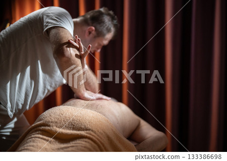 Professional male massage therapist performing deep tissue back massage on an older man lying on a massage table covered with a towel. Relaxing wellness treatment in a modern spa room with warm light Professional male massage therapist performing deep tissue back massage on an older man lying on a massage table covered with a towel. Relaxing wellness treatment in a modern spa room with warm light 133386698