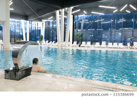 Contemporary indoor swimming pool with clean blue water, stylish lighting, and a metal waterfall fountain surrounded by white lounge chairs. Concept of wellness, luxury, relaxation, and modern spa 133386699