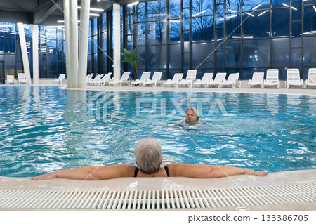 Two senior women enjoying relaxation while leaning on the edge of a modern indoor swimming pool in the evening. Concept of wellness, healthy lifestyle, spa, leisure, and friendship for older adults. Two senior women enjoying relaxation while leaning on the edge of a modern indoor swimming pool in the evening. Concept of wellness, healthy lifestyle, spa, leisure, and friendship for older adults. 133386705