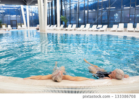 Two senior women enjoying relaxation while leaning on the edge of a modern indoor swimming pool in the evening. Concept of wellness, healthy lifestyle, spa, leisure, and friendship for older adults. 133386708