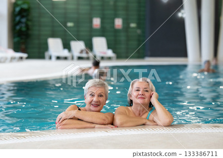 Two smiling senior women enjoying leisure time in an indoor swimming pool, leaning on the poolside. Concept of friendship, wellness, active lifestyle, relaxation, and healthy aging. 133386711
