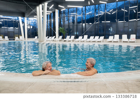 Two smiling senior women enjoying leisure time in an indoor swimming pool, leaning on the poolside. Concept of friendship, wellness, active lifestyle, relaxation, and healthy aging. 133386718