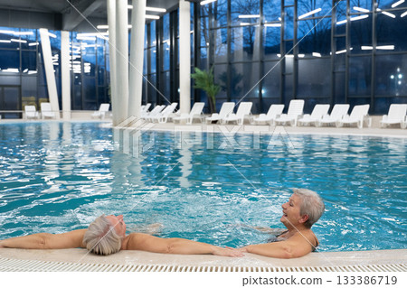 Two smiling senior women enjoying leisure time in an indoor swimming pool, leaning on the poolside. Concept of friendship, wellness, active lifestyle, relaxation, and healthy aging. 133386719