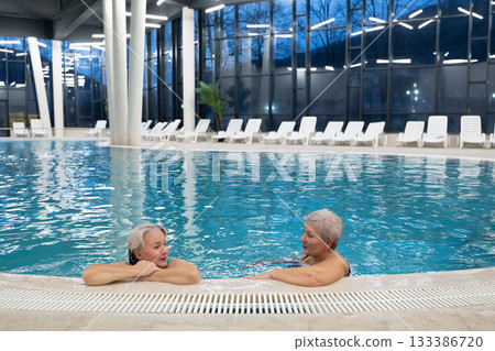 Two smiling senior women enjoying leisure time in an indoor swimming pool, leaning on the poolside. Concept of friendship, wellness, active lifestyle, relaxation, and healthy aging. 133386720