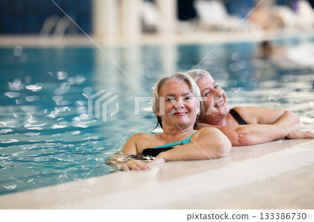 Two smiling senior women enjoying leisure time in an indoor swimming pool, leaning on the poolside. Concept of friendship, wellness, active lifestyle, relaxation, and healthy aging. Two smiling senior women enjoying leisure time in an indoor swimming pool, leaning on the poolside. Concept of friendship, wellness, active lifestyle, relaxation, and healthy aging. 133386730