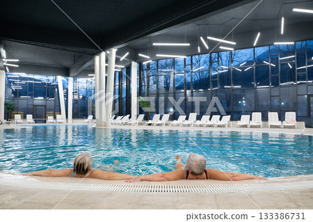 Two senior women enjoying relaxation while leaning on the edge of a modern indoor swimming pool in the evening. Concept of wellness, healthy lifestyle, spa, leisure, and friendship for older adults. 133386731