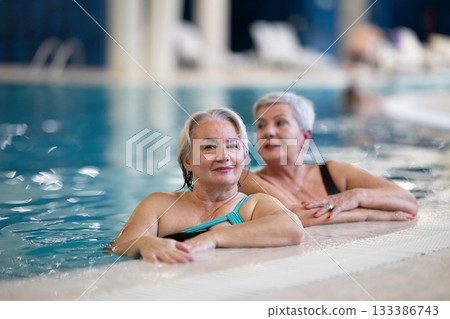 Two smiling senior women enjoying leisure time in an indoor swimming pool, leaning on the poolside. Concept of friendship, wellness, active lifestyle, relaxation, and healthy aging. 133386743