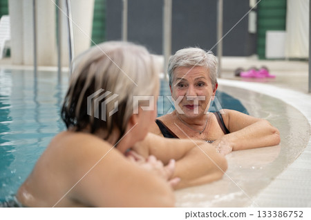 Two smiling senior women enjoying leisure time in an indoor swimming pool, leaning on the poolside. Concept of friendship, wellness, active lifestyle, relaxation, and healthy aging. Two smiling senior women enjoying leisure time in an indoor swimming pool, leaning on the poolside. Concept of friendship, wellness, active lifestyle, relaxation, and healthy aging. 133386752