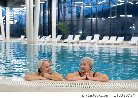 Two smiling senior women enjoying leisure time in an indoor swimming pool, leaning on the poolside. Concept of friendship, wellness, active lifestyle, relaxation, and healthy aging. Two smiling senior women enjoying leisure time in an indoor swimming pool, leaning on the poolside. Concept of friendship, wellness, active lifestyle, relaxation, and healthy aging. 133386754
