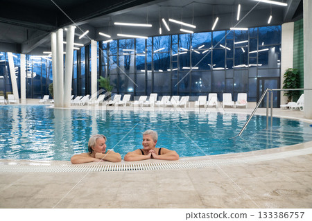 Two smiling senior women enjoying leisure time in an indoor swimming pool, leaning on the poolside. Concept of friendship, wellness, active lifestyle, relaxation, and healthy aging. Two smiling senior women enjoying leisure time in an indoor swimming pool, leaning on the poolside. Concept of friendship, wellness, active lifestyle, relaxation, and healthy aging. 133386757