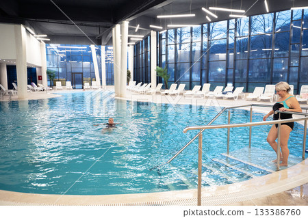 Elderly woman sitting by the edge of a modern indoor spa pool, enjoying the relaxing water jets. Wellness and relaxation concept in a luxurious spa or wellness center. 133386760