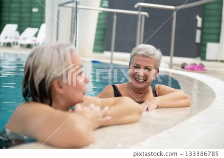 Two smiling senior women enjoying leisure time in an indoor swimming pool, leaning on the poolside. Concept of friendship, wellness, active lifestyle, relaxation, and healthy aging. 133386785