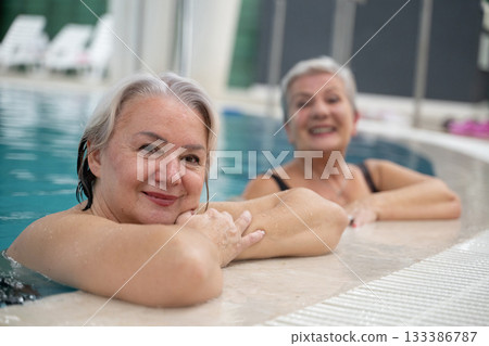 Two smiling senior women enjoying leisure time in an indoor swimming pool, leaning on the poolside. Concept of friendship, wellness, active lifestyle, relaxation, and healthy aging. 133386787