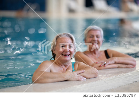 Two smiling senior women enjoying leisure time in an indoor swimming pool, leaning on the poolside. Concept of friendship, wellness, active lifestyle, relaxation, and healthy aging. Two smiling senior women enjoying leisure time in an indoor swimming pool, leaning on the poolside. Concept of friendship, wellness, active lifestyle, relaxation, and healthy aging. 133386797